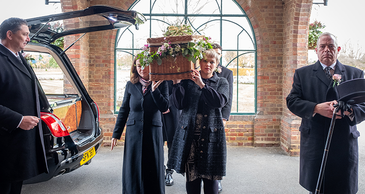 Four people carrying a flower-laden wicker coffin into a chapel with two funeral director staff either side and a hearse 